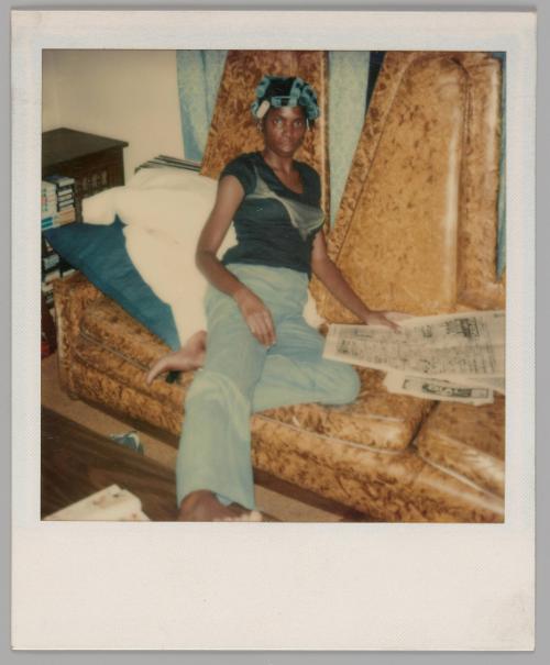 [Woman with blue curlers in her hair and flared jeans sitting with the newspaper on an orange couch, there are white a blue pillows behind her]