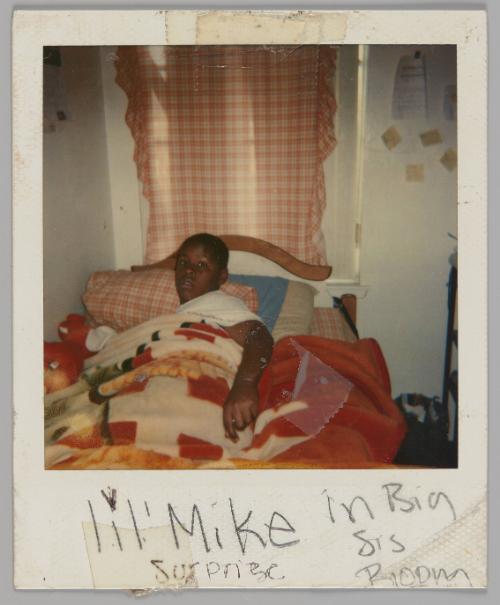 lil' Mike in Big Sis Room Surprize [Boy wearing white t-shirt in bed under quilt and looking up at camera, gingham curtain covering window behind bed]