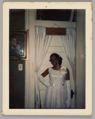 [Teenage girl in white dress, evening gloves and corsage posing with one hand on hip in front of white door with curtain, portrait of Jackie Onassis and J.F.K. on wall]