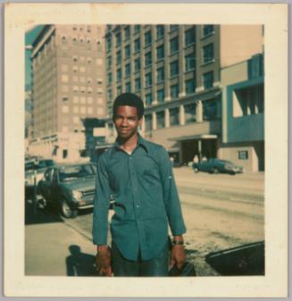 [Young man in blue dress shirt standing on city sidewalk, parked car behind him and buildings across the street]