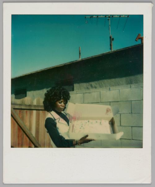 [Woman in black shirt with white vest standing outside and holding open a cake box]