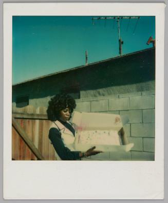 [Woman in black shirt with white vest standing outside and holding open a cake box]