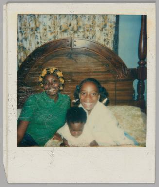 [Young woman with yellow curlers in her hair and wearing a green polo shirt sitting on a bed with an older girl and baby, the baby is looking down]