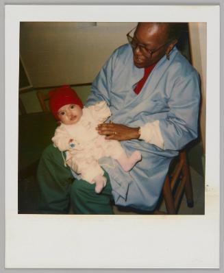 [Woman in glasses, blue jacket and green pants holding a baby in a red hat on her lap]
