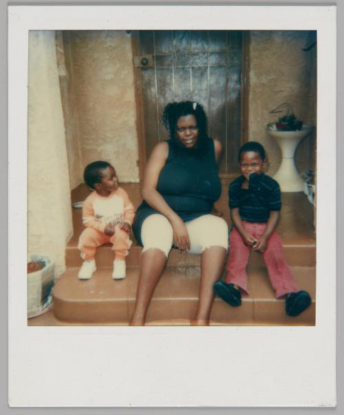 [Woman sitting between two boys on the steps outside a wood door, boy on right is wearing red pants and sticking his legs straight out]