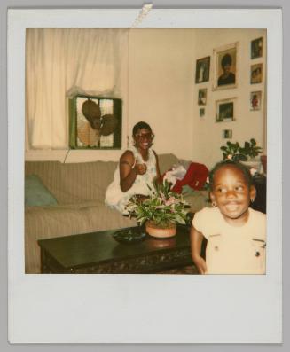 [Woman in glasses smiling and sitting on couch with a girl standing close to camera and smiling, box fan in the window and family portraits on the wall]