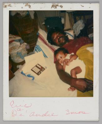 Eric + De'Andre 3 mos [Man in red t-shirt lying down with his arm around a baby dressed in white, toy phone and laundry basket to the left]