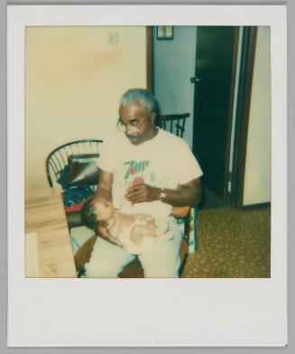 [Man with glasses in 7-Up t-shirt sitting down and feeding a bottle to a baby]