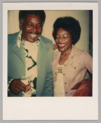 [Woman in studded pink jacket laughing and posing beside a man with a moustache and a drink can in his hand]