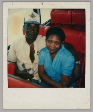 [Man in white shirt and baseball cap with his arm around a woman in a blue short-sleeved shirt, they are sitting on red bus seats]