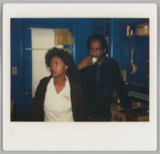 [Woman in white shirt and cardigan with name tag standing beside man sipping from a small cup, they are in a blue kitchen]