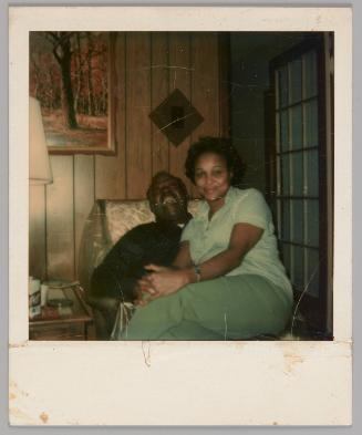 [Woman in green pants sitting on man's lap, he is in an armchair laughing, they are in a wood panelled room]