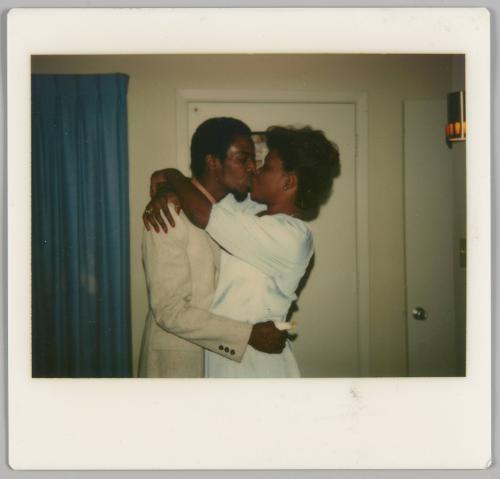 [Man in light-coloured suit kissing woman in white dress, they are inside and there is a blue curtain and door behind them]