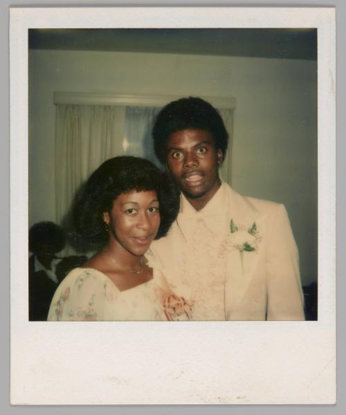 [Woman in white dress with pink corsage standing with a man in a light-coloured tuxedo and a white boutonnière, there is a window behind them]