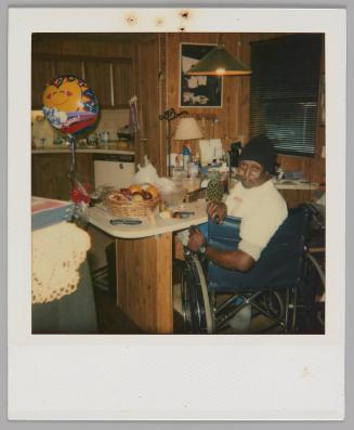 [Man in black hat sitting at a table in a wheelchair, he is turning back to smile at the camera, there is a bowl of fruit on the table and a balloon to the left]