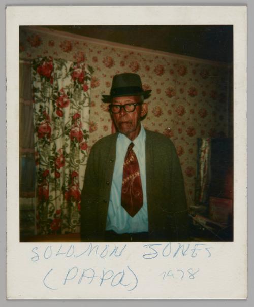 Solomon Jones (Papa) 1978 [Older man in fedora hat, black rimmed glasses, a blue shirt and red tiestading in a room with floral wallpaper and curtains]