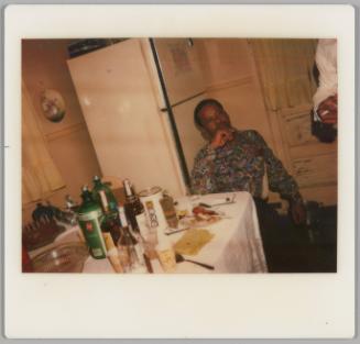 [Man in patterned shirt sitting with his elbow on a kitchen table and his hand on his chin, there is a refrigerator behind him]