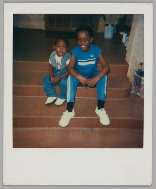 [Two kids sitting on steps, boy on right wearing blue t-shirt and track pants with white stripes and white velcro shoes]