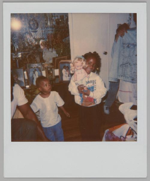 [Girl in sweatshirt holding up a doll with blonde hair with young boy on the left, there is a dresser with photographs behind them]