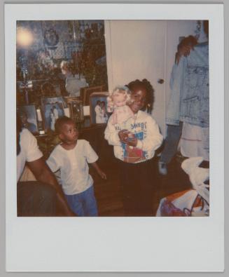 [Girl in sweatshirt holding up a doll with blonde hair with young boy on the left, there is a dresser with photographs behind them]