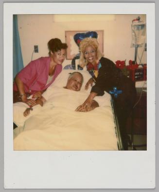 [Man lying down in hospital bed with two women on either side of him, woman on left in pink shirt and woman on right is blonde]