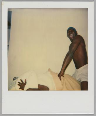 [Man in shorts laughing and jumping on someone in bed, they are under the covers holding a pillow over their head]