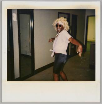 [Man in blonde curly wig, sunglasses, and pink shirt tied in the front spreading his arms wide and smiling at camera]