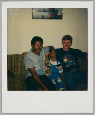 [Two men sitting on couch with toddler between them, toddler is holding two polaroids and a teddy bear, the man to the left is covering their face]