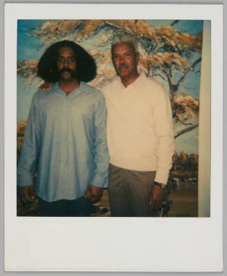 [Two men standing together in front of a mural of an outdoor scene, man on left has shoulder-length hair and a blue dress shirt]