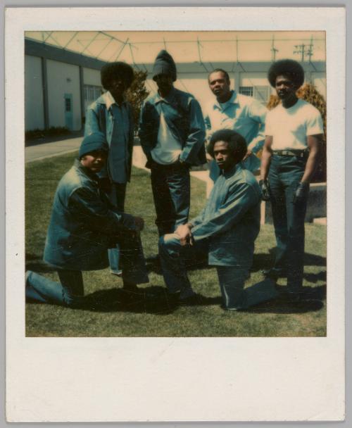 [Six men in fenced in lawn, four men standing and two men in front wearing jackets and kneeling with both their hands on their knees]