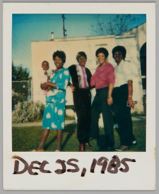 Dec 25, 1985 [Four women standing in a row outside, woman on left holds baby and is wearing a blue and white dress]