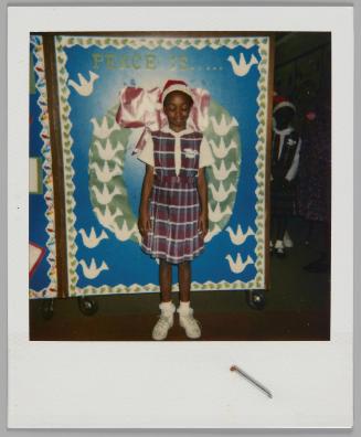 [Girl in plaid uniform and Santa Claus hat standing in front of poster that reads "Peace Is....." and picture of a wreath with doves]
