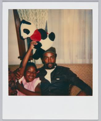 [Man sitting next to little girl in pink who is holding up panda plushed animal]
