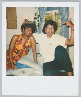 [Two woman sitting on bed smiling, woman on left wears a yellow and red floral ensemble]