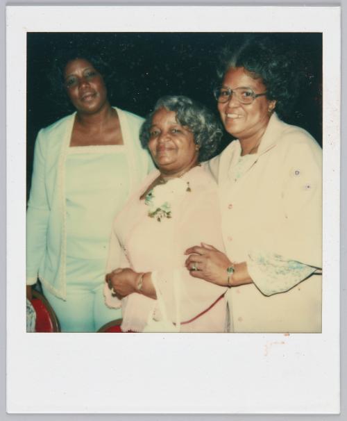 [Three women in pastel coloured outfits smiling, woman in middle has flower corsage pinned to her chest]