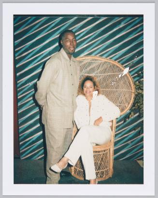 [Young man in light brown outfit standing next to woman in white outfit sitting on larger wicker chair]