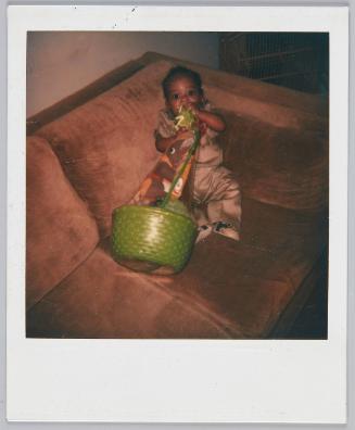 [Infant propped up in couch corner sucking on wrapping of gift basket]