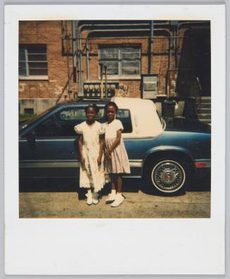 [Two little girls in formal dresses posing in front of car, brick building behind them]