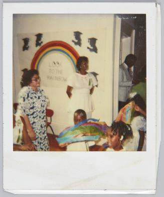 [Woman wearing white dress and ribbon on shoulder standing with hands behind back, sign that reads 'Look To The Rainbow' hung on wall]