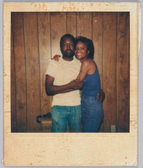 [Woman in blue tank top hugging man in white tshirt, both standing in front of wood paneled wall]