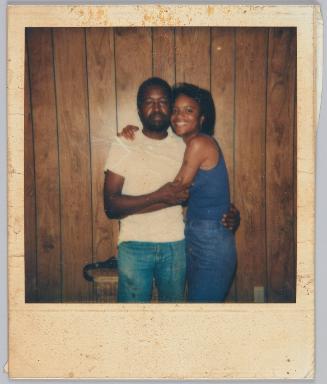 [Woman in blue tank top hugging man in white tshirt, both standing in front of wood paneled wall]