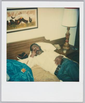 [Man with moustache fully clothed and asleep in bed]