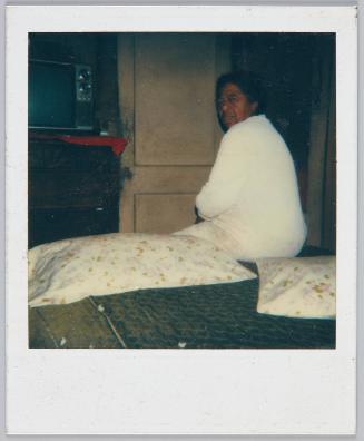 [Elderly woman sitting on edge of bed, two pillows with floral patterns next to her]