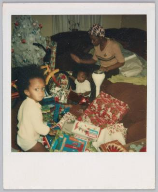 [Toddler and infant surrounded by Christmas gifts, young woman sitting on couch behind them]