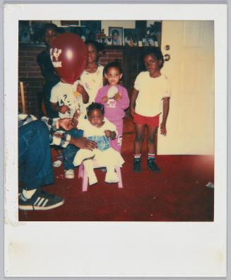 [Group of five children standing around toddler girl sitting in little plastic pink chair]