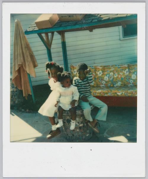 [Three kids sitting on concrete post, little girl on left wears a white dress and red hair accessories]