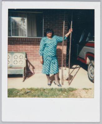 [Woman wearing large sunglasses and turquoise patterned outfit poses on porch of house]