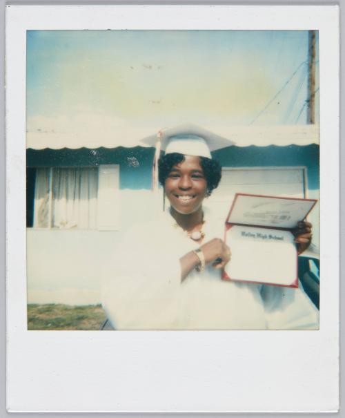 [Adolescent girl wearing white graduation cap and gown smiles while holding up high school diploma]