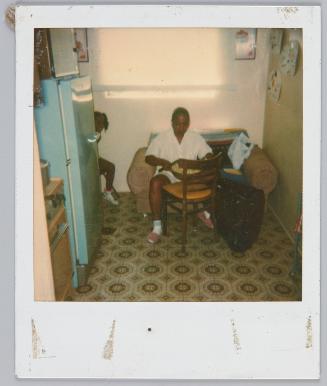[Woman sitting on seat in kitchen holding a bowl, little girl peeking out behind refrigerator]