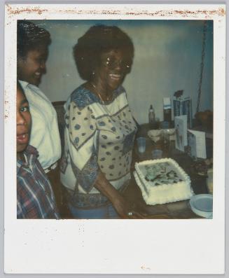 [Older woman wearing large-rimmed glasses and patterned top standing in front of cake]